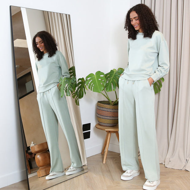 Woman in a light green outfit standing next to a mirror in a room with a plant.