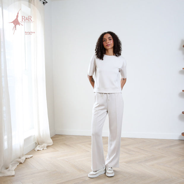 Woman in white outfit standing in a minimalistic room with 'R&R' logo in the corner.