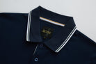Close-up of the 6th Sense Astound Eclipse Polo in Eclipse, showing the ribbed collar with double white stripes, buttoned placket, and inner neck tape with gold and white stripes. A black 6th Sense Global Designs label with a size M tag is visible at the back neck.