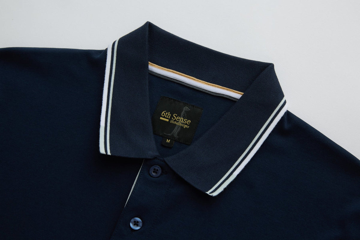 Close-up of the 6th Sense Astound Eclipse Polo in Eclipse, showing the ribbed collar with double white stripes, buttoned placket, and inner neck tape with gold and white stripes. A black 6th Sense Global Designs label with a size M tag is visible at the back neck.