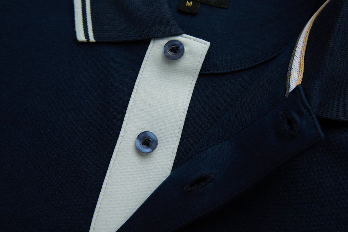 Close-up of the 6th Sense Astound Eclipse Polo placket in Eclipse, showing two dark buttons on a light placket, a striped collar tip, a small M size label, and contrast trim along the inner neckline.