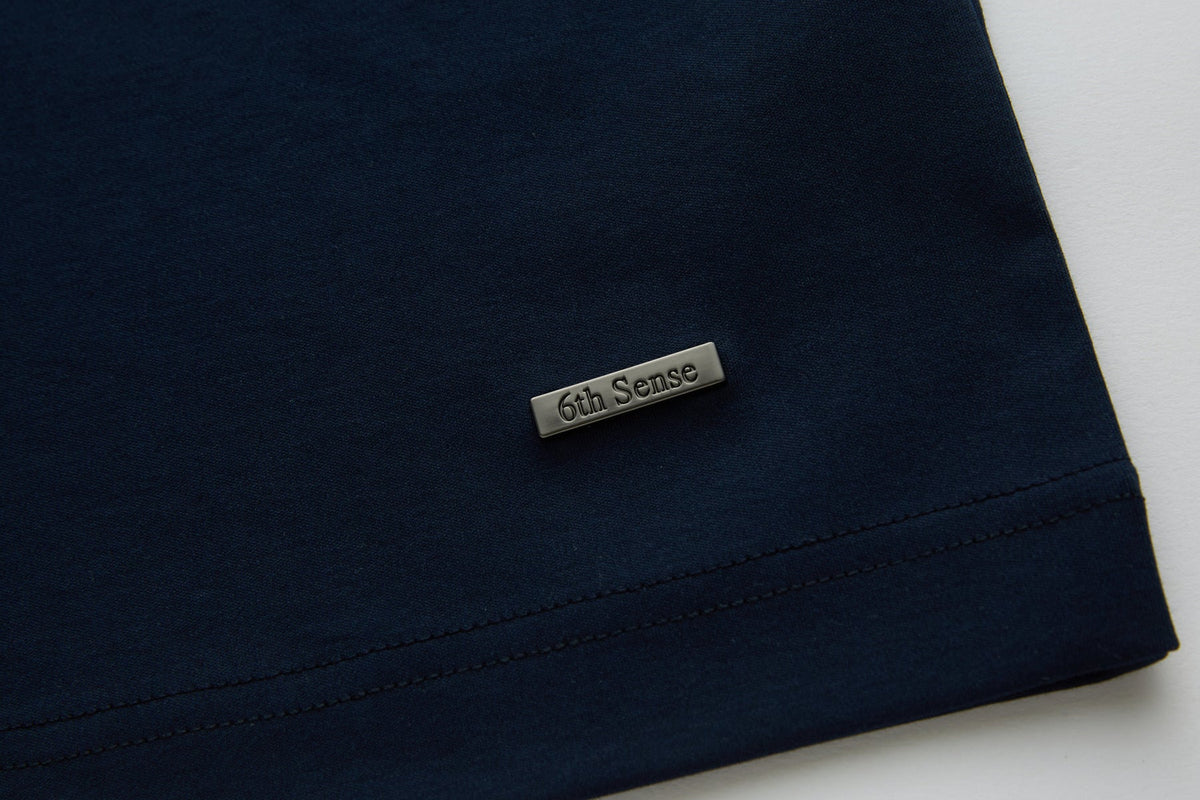 Close-up of the lower hem on the 6th Sense Astound Eclipse Polo in Eclipse, showing dark fabric with double stitching and a small rectangular metal tag engraved with the 6th Sense logo.