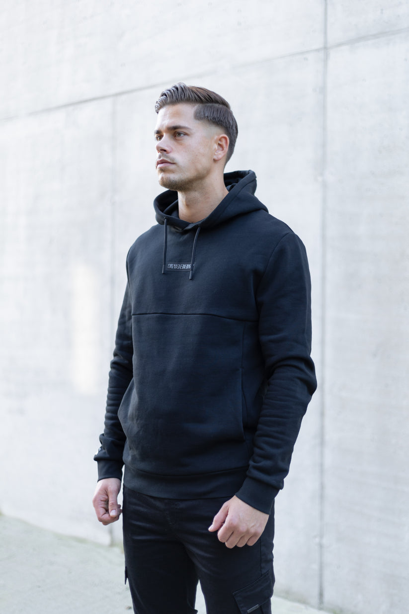 Man wearing a black hoodie with a brand logo against a light gray wall.