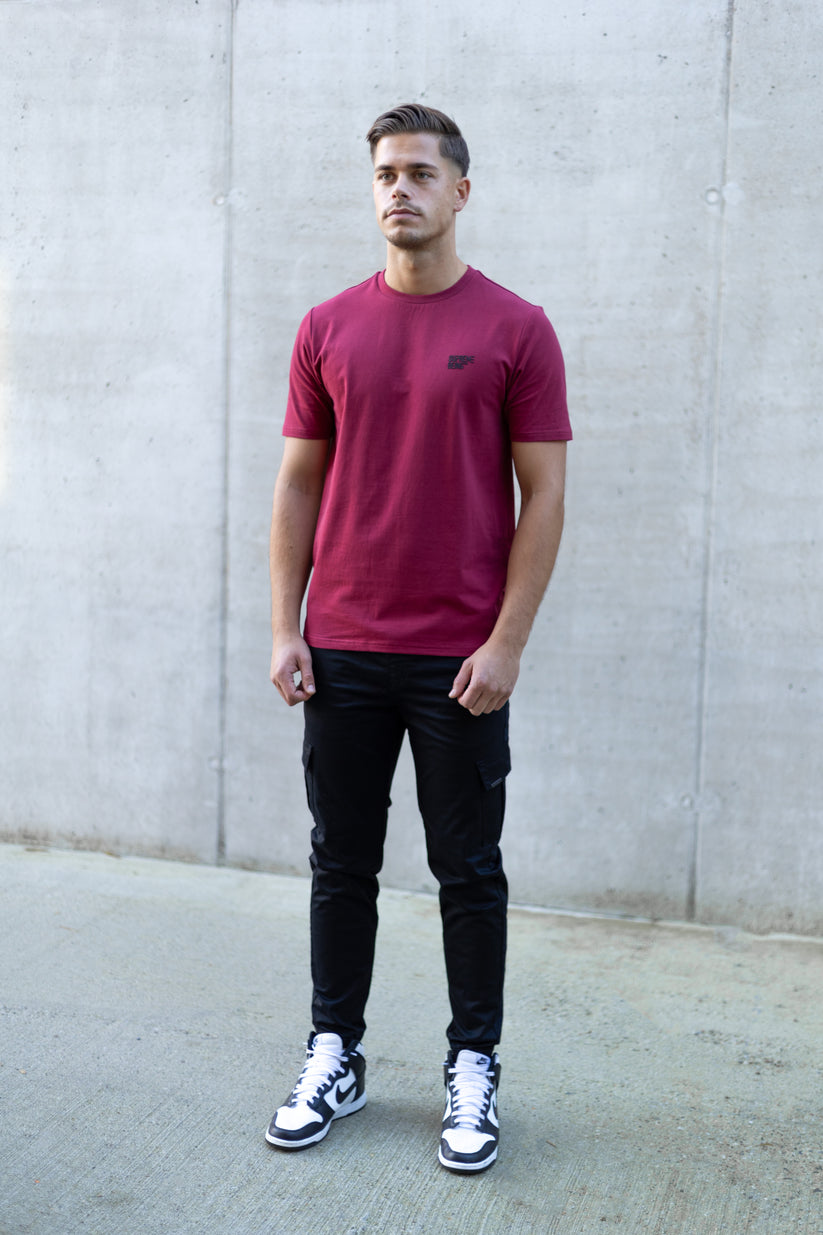 Man wearing a red t-shirt, black pants, and white sneakers against a gray concrete wall.