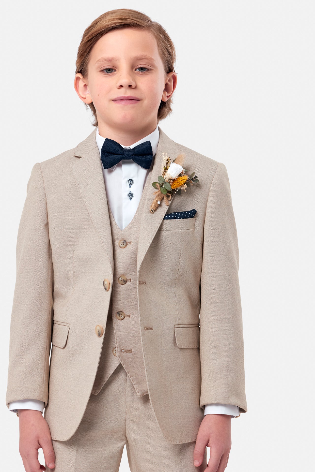 Young boy wearing a beige suit with a white shirt, navy bow tie, and floral boutonniere on a light gray background.