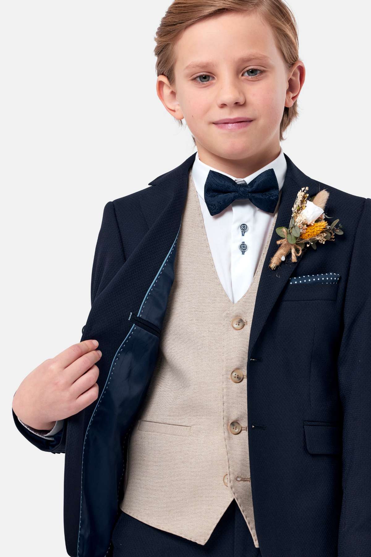 Young boy in a formal navy suit with a bow tie, stone waistcoat and boutonniere on a light gray background