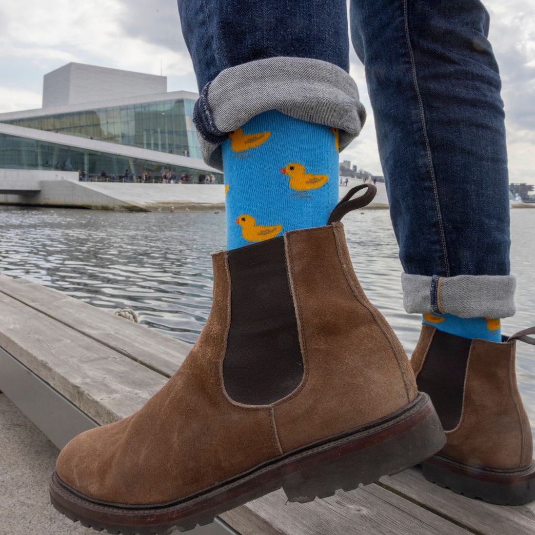 Person wearing brown boots and blue socks with yellow duck pattern, standing on a wooden platform by water.