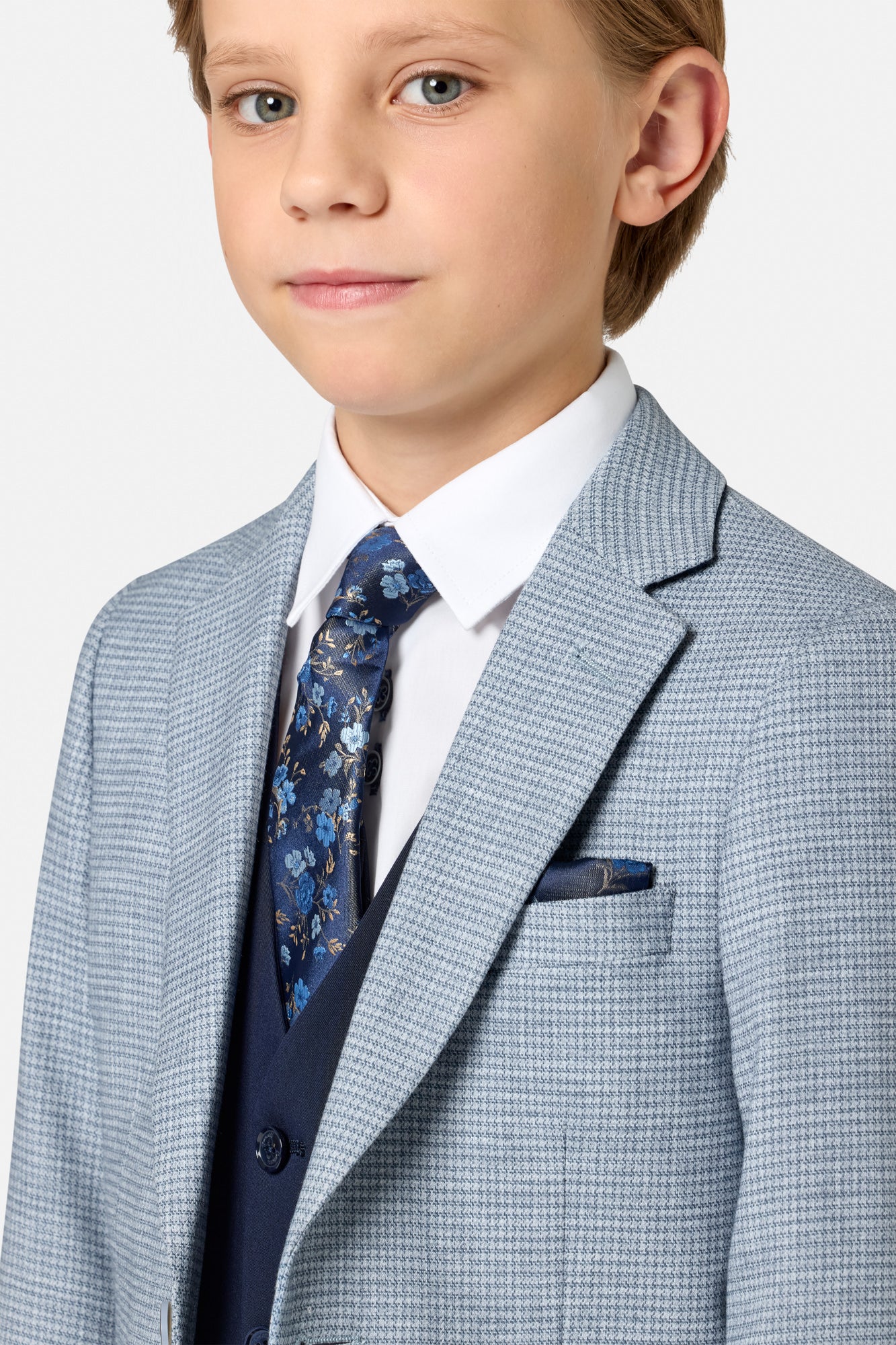 Young boy wearing a light blue checkered suit jacket with a white shirt and patterned tie on a plain background