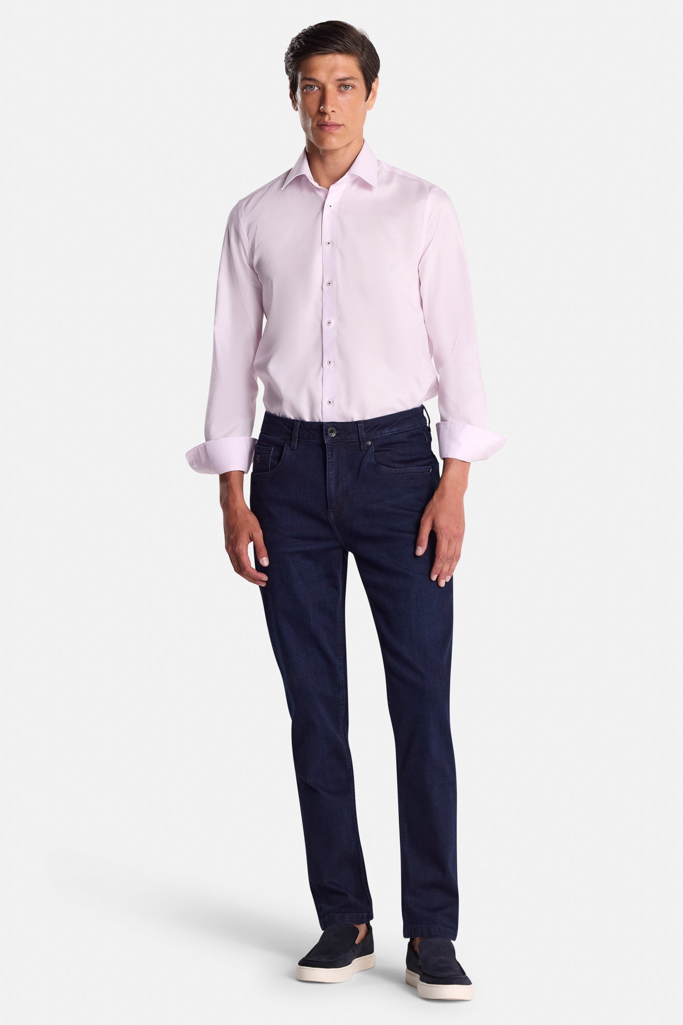Man wearing a light pink dress shirt and dark blue jeans on a white background