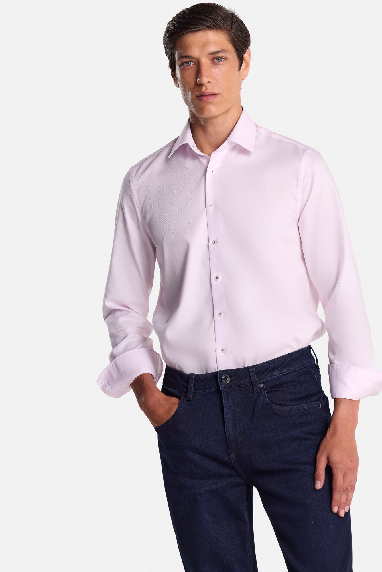 Man wearing a light pink dress shirt and dark blue jeans on a white background