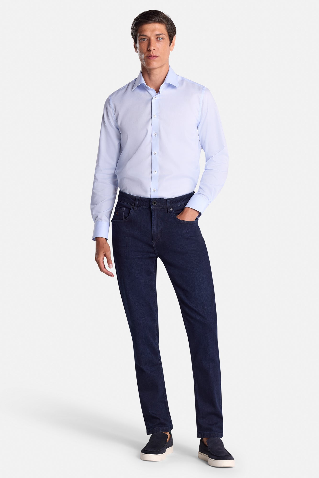 Man wearing a light blue dress shirt and dark blue jeans on a white background