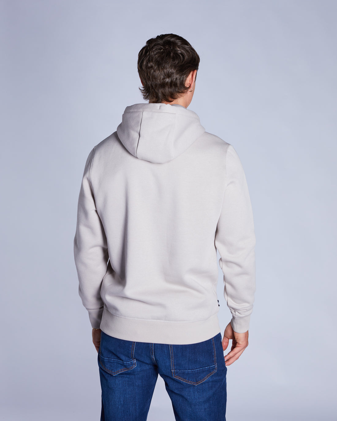 Person wearing a beige  hoodie and blue jeans against a plain background
