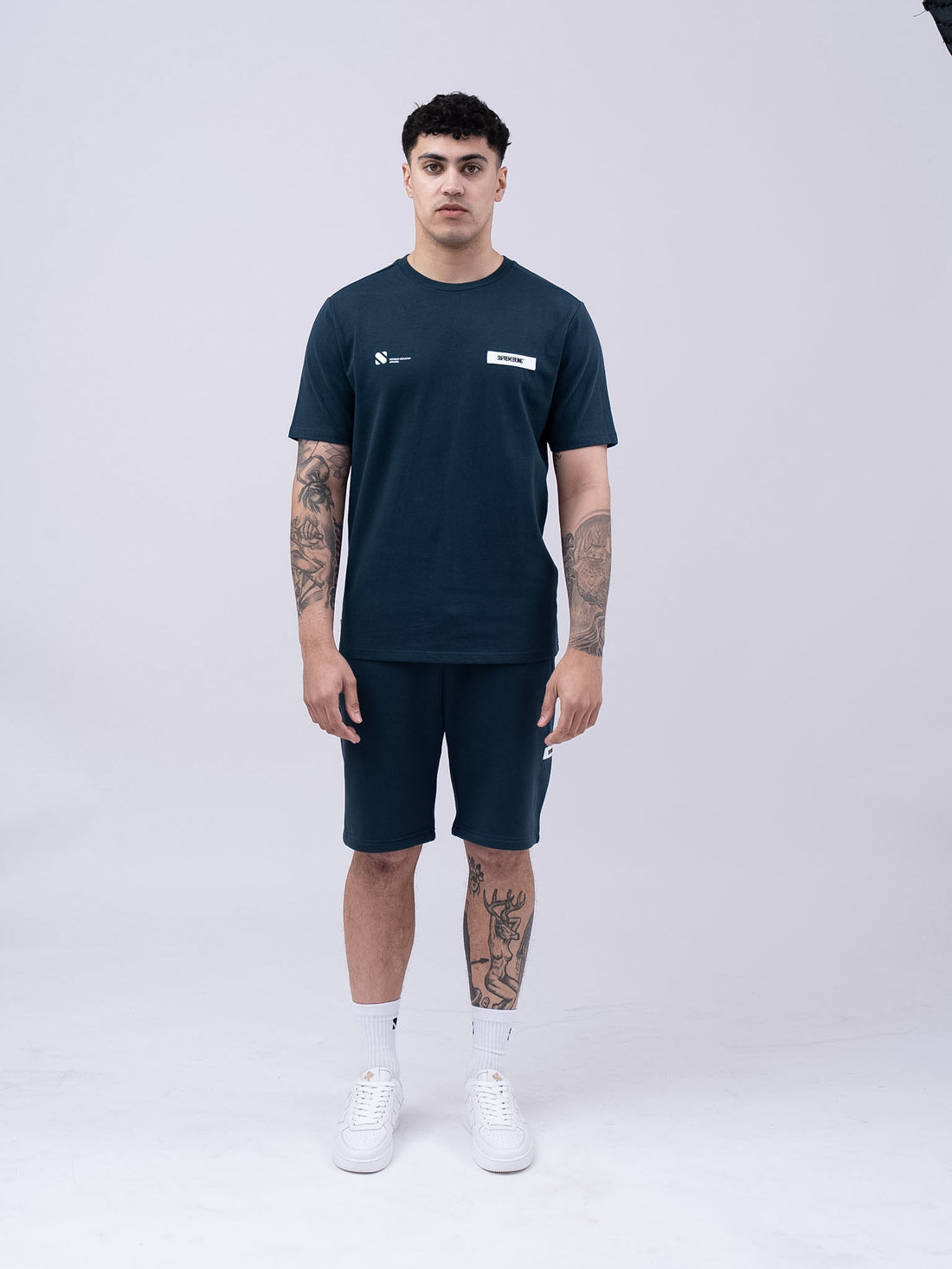 Man wearing a navy t-shirt and shorts with visible tattoos on a white background