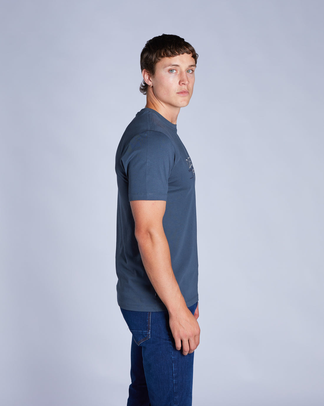 Man wearing a petrol blue tee and blue jeans side view looking at camera
