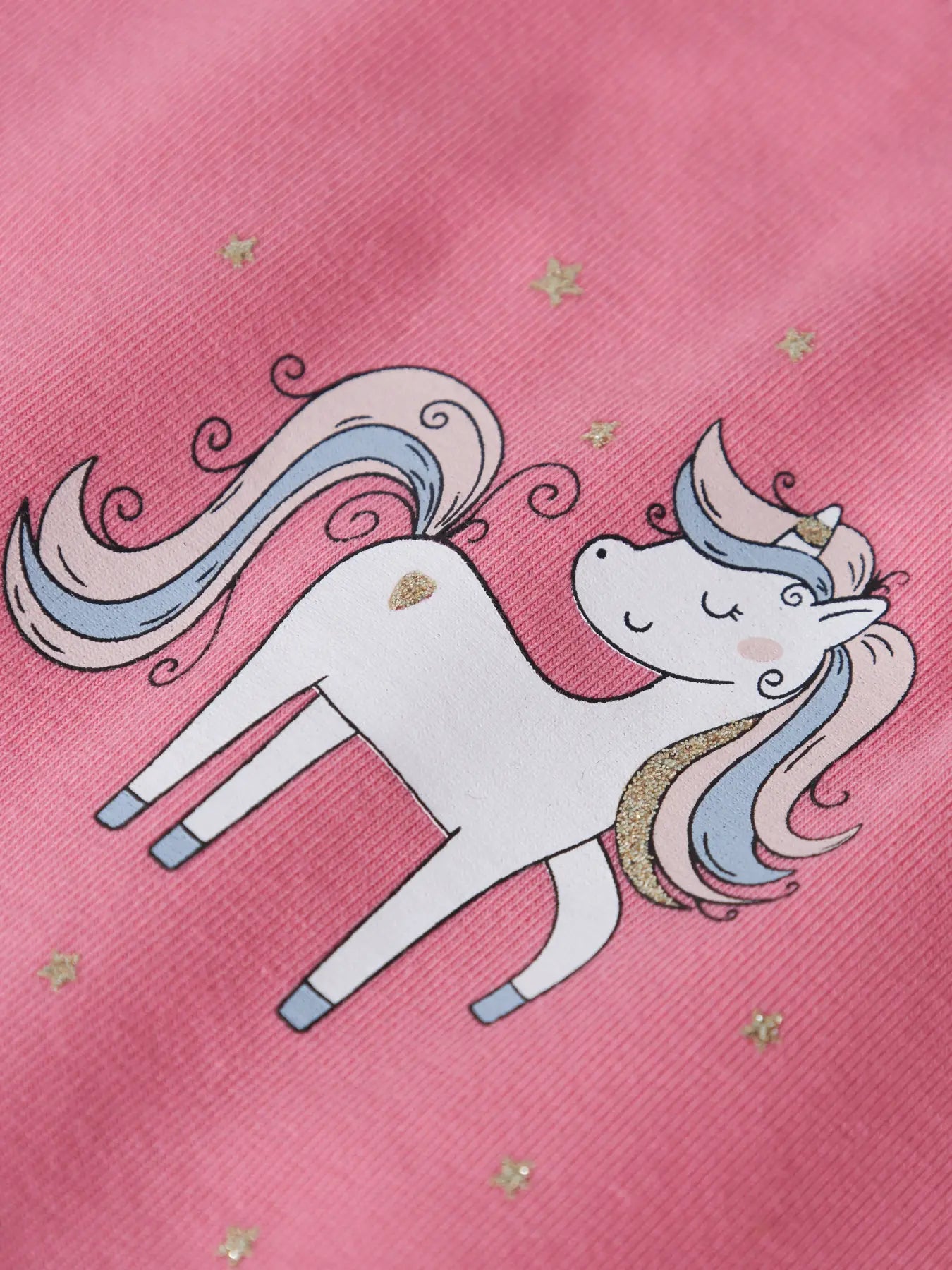 A close-up of a pink fabric featuring a cartoon unicorn graphic with pastel blue and pink hair, gold stars, and a small gold heart on the side.