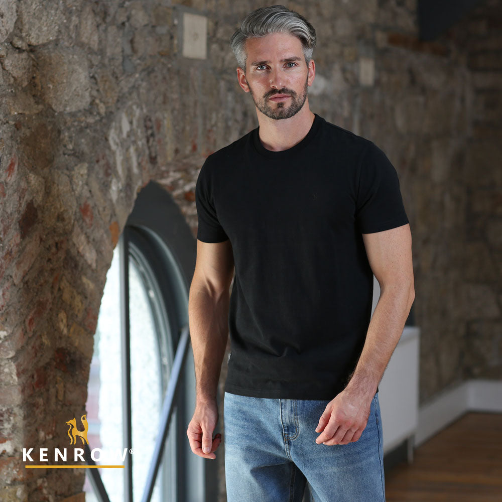 Man wearing a black t-shirt and blue jeans standing against a stone wall.