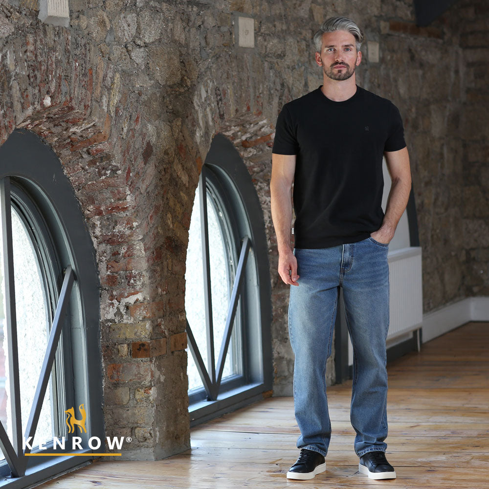 Man wearing a black t-shirt and blue jeans standing in a room with stone walls and large windows.