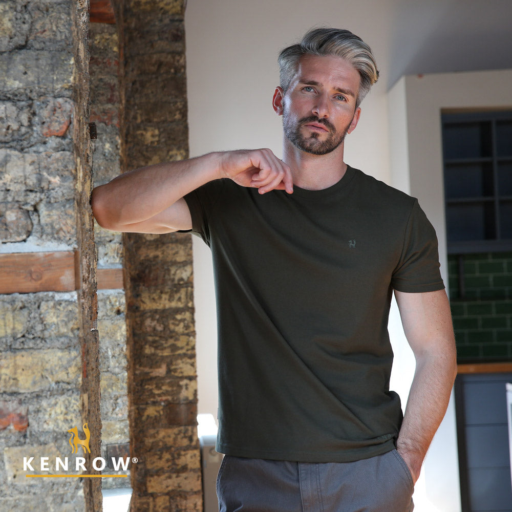 Man wearing a dark green t-shirt standing indoors with stone wall background