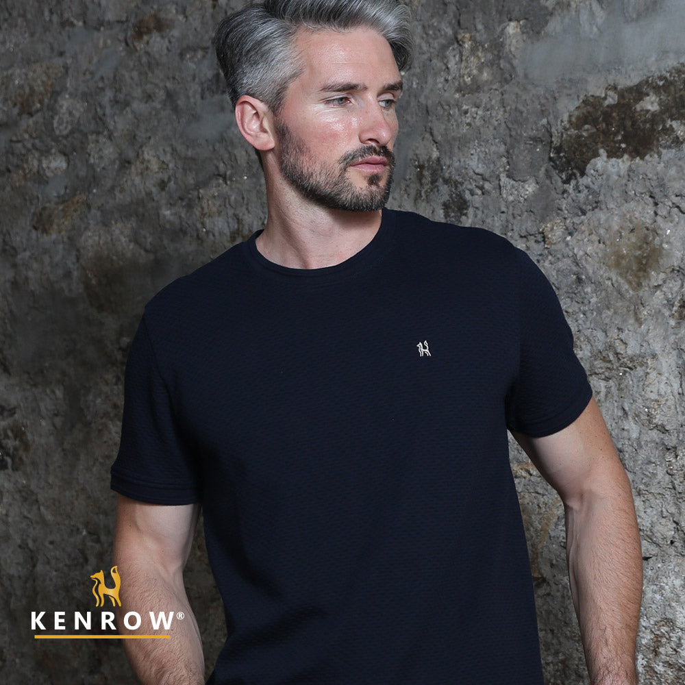 Man wearing a black t-shirt with a small logo against a textured wall, featuring the brand 'Kenrow'.