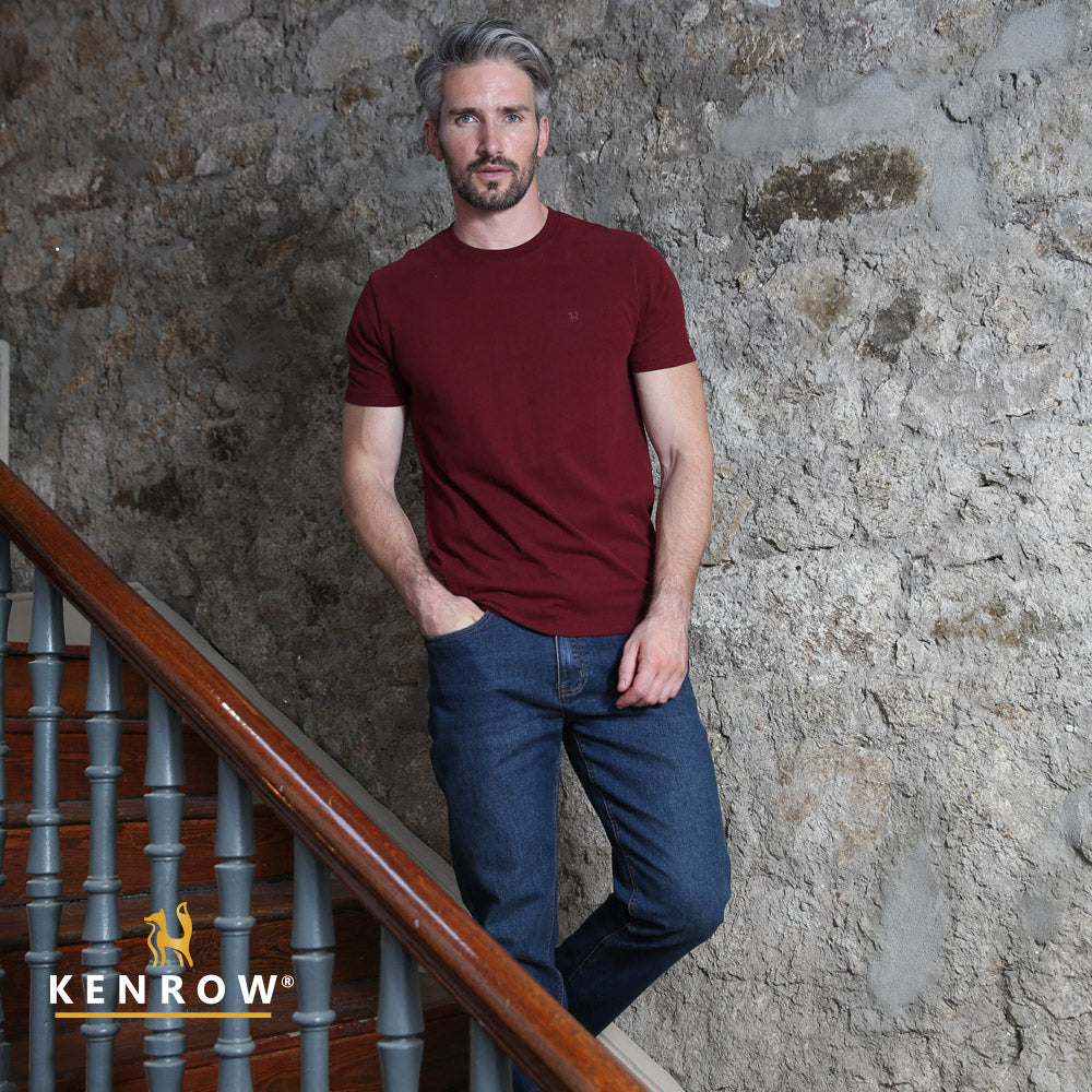 Man wearing a red t-shirt and blue jeans leaning against a stone wall with 'Kenrow' branding.