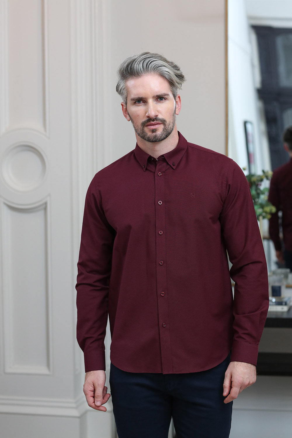 A man with styled gray hair and a beard wearing a long-sleeve burgundy button-down shirt and dark trousers, standing in an indoor setting with a white paneled wall on the left.