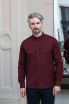 A man with styled gray hair and a beard wearing a long-sleeve burgundy button-down shirt and dark trousers, standing in an indoor setting with a white paneled wall on the left.