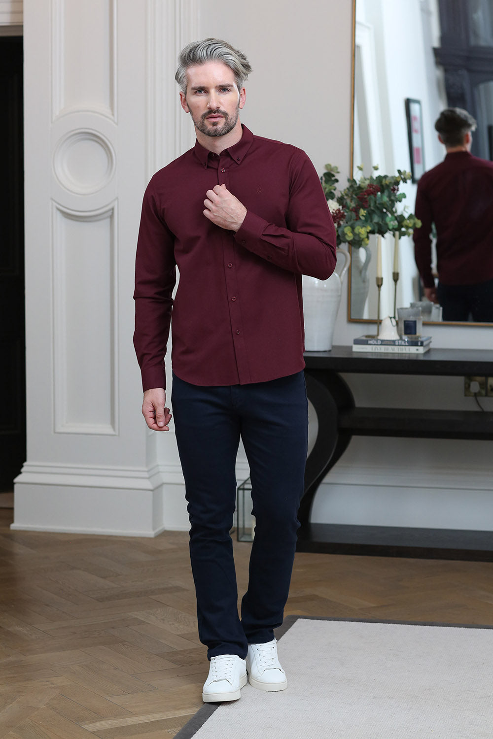 A man wearing a long-sleeve burgundy button-down shirt, dark navy pants, and white sneakers, standing in a modern indoor setting with a mirror reflecting his back.