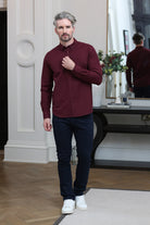 A man wearing a long-sleeve burgundy button-down shirt, dark navy pants, and white sneakers, standing in a modern indoor setting with a mirror reflecting his back.