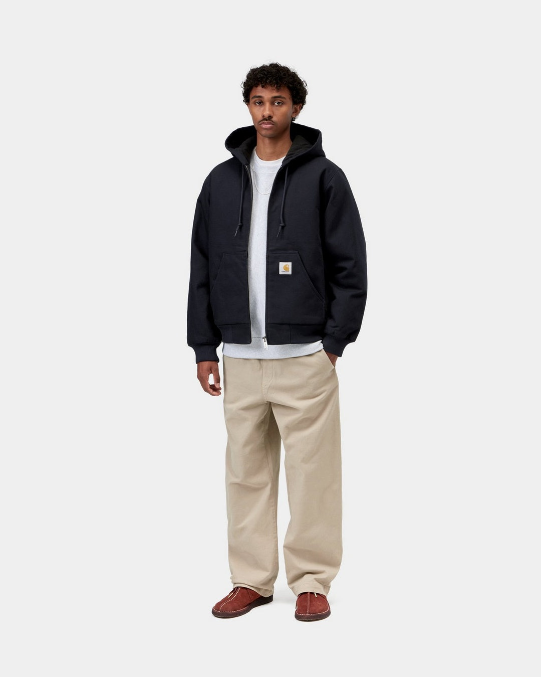 Carhartt WIP Active Jacket in Dark Navy, front view, worn by a model with a white T-shirt and beige trousers. The jacket is zipped with the hood down, showing drawstrings, front pockets, ribbed cuffs and hem, and a square Carhartt label on the left pocket.
