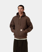 Front view of the Carhartt WIP Active Jacket in Tobacco, worn with the hood down and hands in the front pockets. The brown jacket features a gold-tone zip, drawstring hood, ribbed cuffs and hem, and a square Carhartt WIP logo patch on the kangaroo pocket.