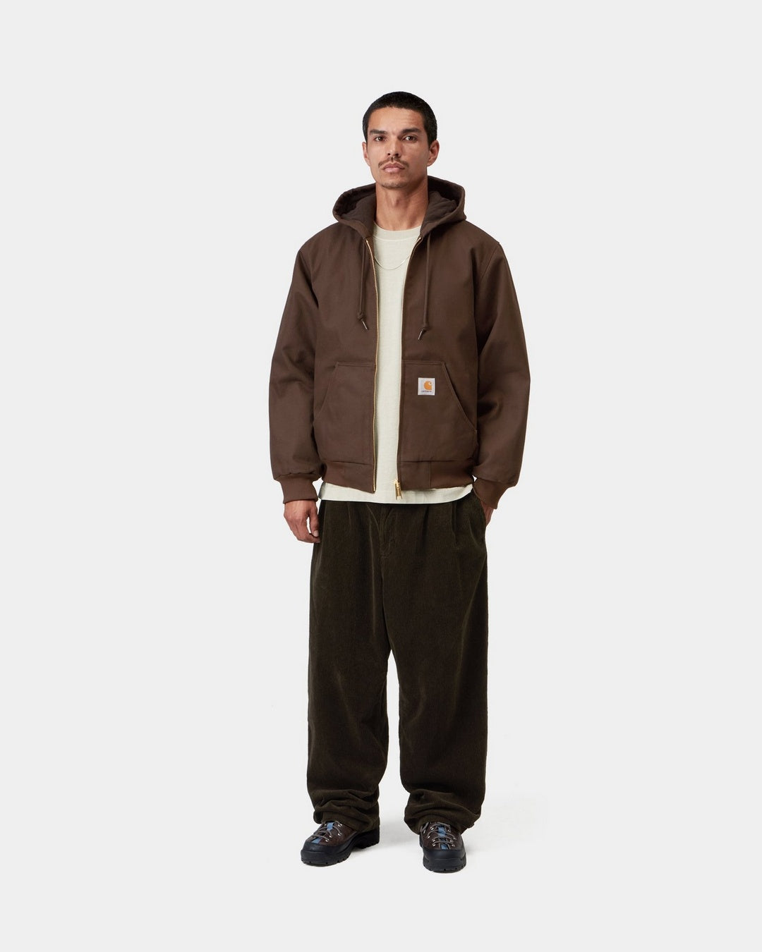 Carhartt WIP Active Jacket in Tobacco worn by a model, front view. The hooded zip jacket is shown unzipped with a white t-shirt underneath, featuring two front pockets, a woven Carhartt WIP label on the left pocket, ribbed cuffs and hem, and an adjustable drawstring hood.
