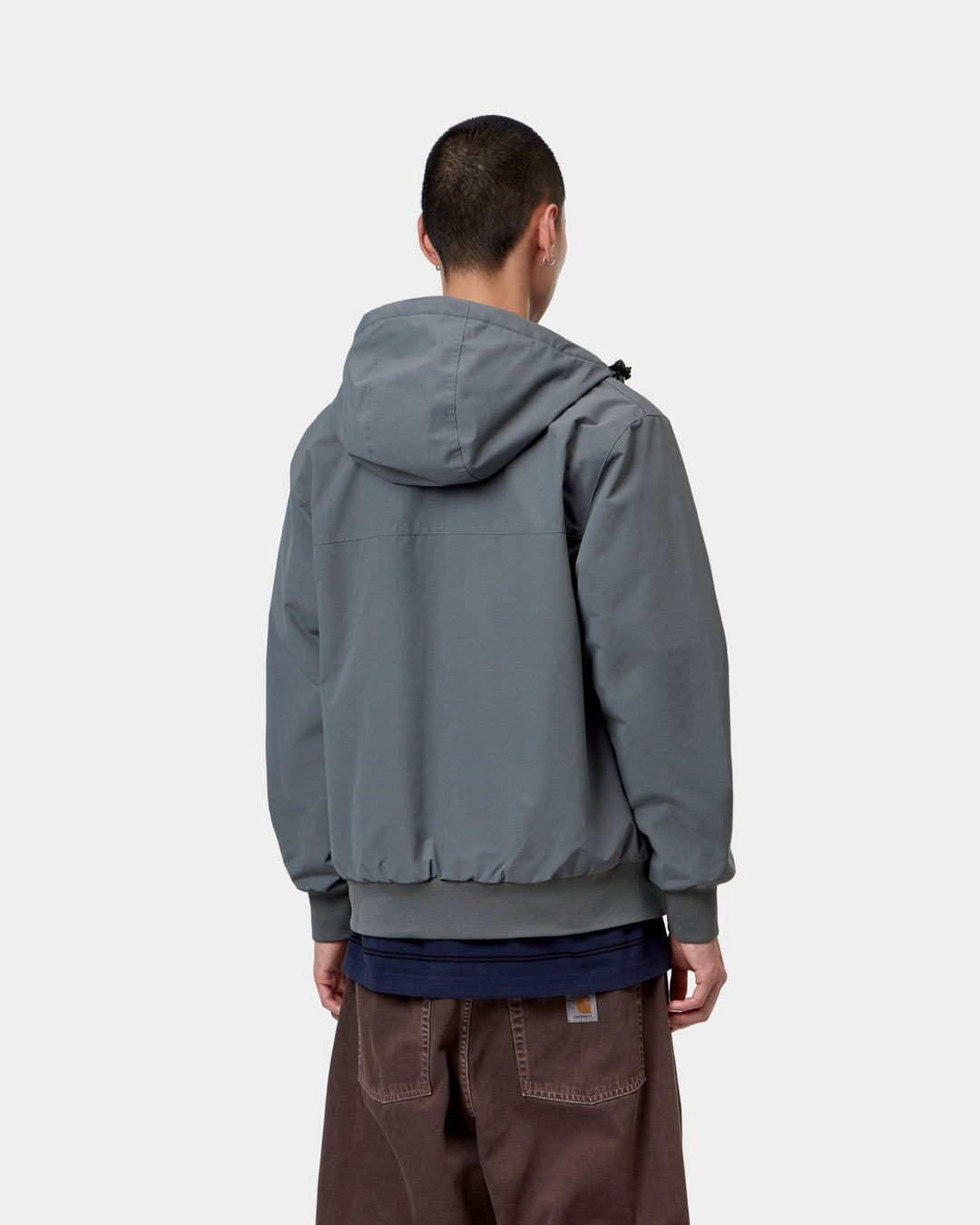 Back view of the Carhartt WIP Hooded Sail Jacket in Styx / White, showing the attached hood, back yoke seam, rib-knit hem and cuffs, and a model wearing it over a dark layer and brown pants.