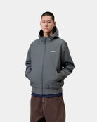 Model wearing Carhartt WIP Hooded Sail Jacket in Styx / White, front view, hands in pockets, hood up, white Carhartt logo on chest.