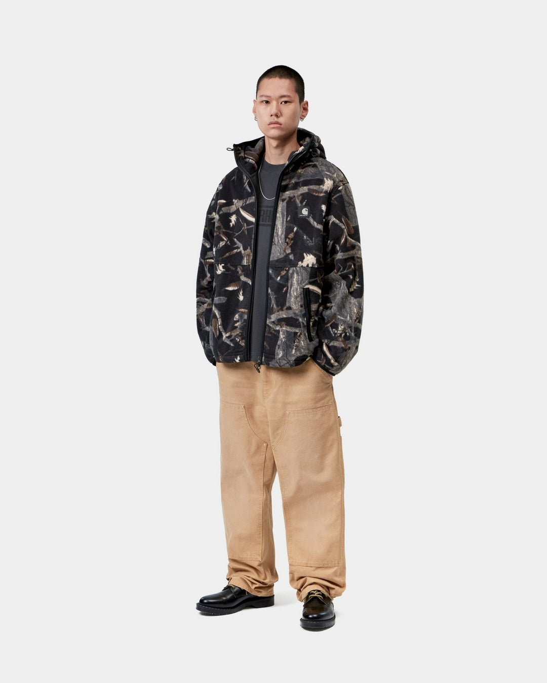 Carhartt WIP Blevin Liner in Camo Feather Tree, Black worn by model, front view, with hood and reflective Square Label visible.