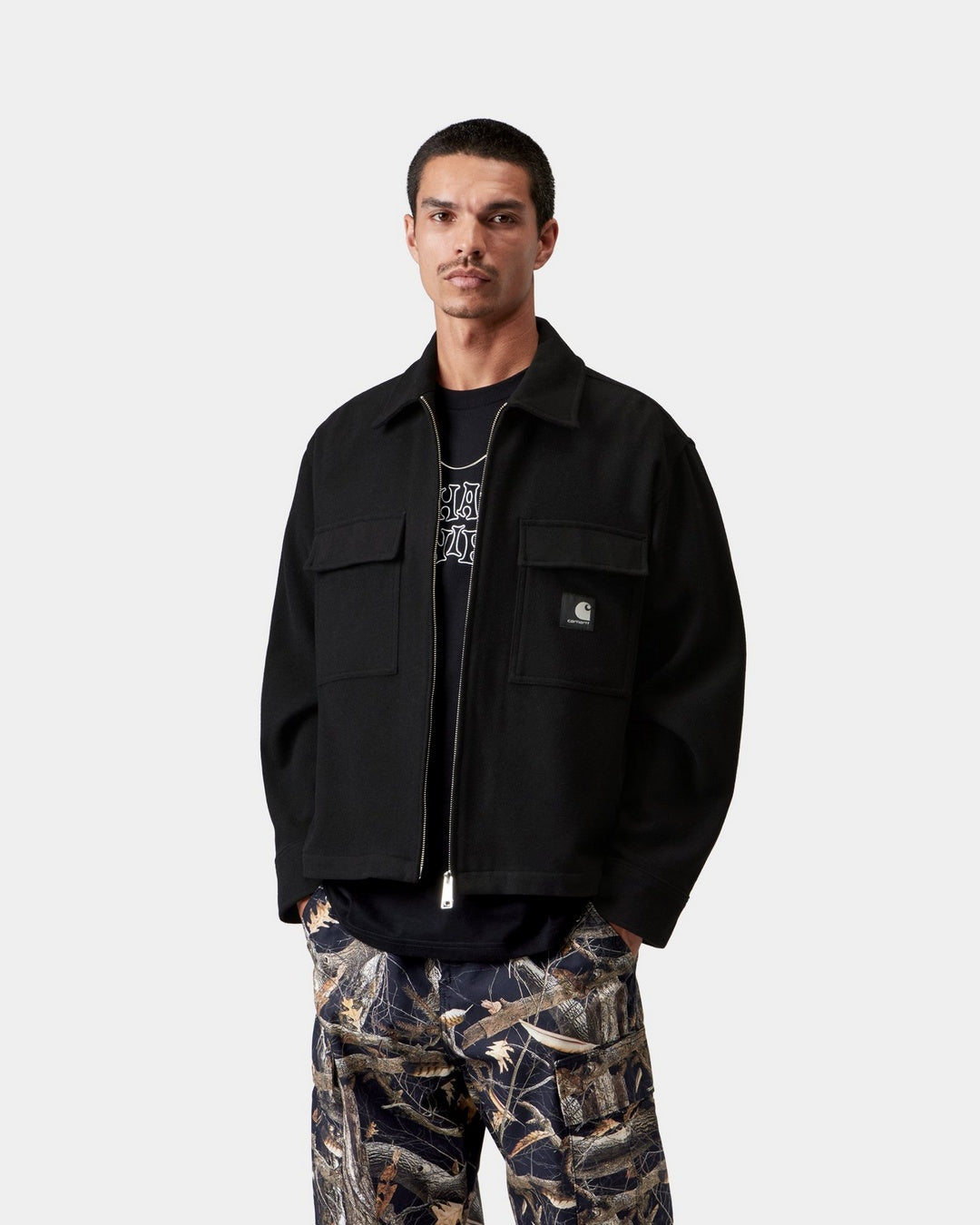 Carhartt WIP Atoka Shirt Jac in Black, front view, worn open over a graphic t-shirt. The black shirt jacket features a pointed collar, silver two-way zip, two chest flap pockets, and a black Square Label on the left pocket.