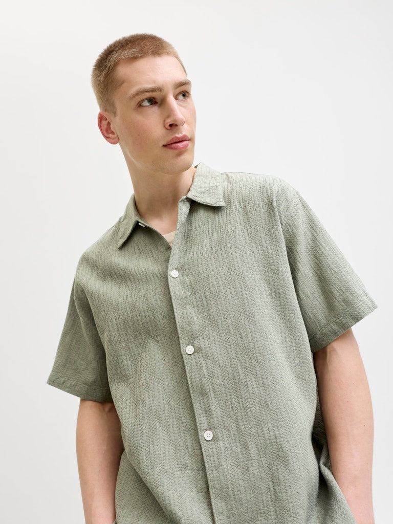 Man wearing montauk wavy texture short sleeve vetiver button-up shirt against a plain background