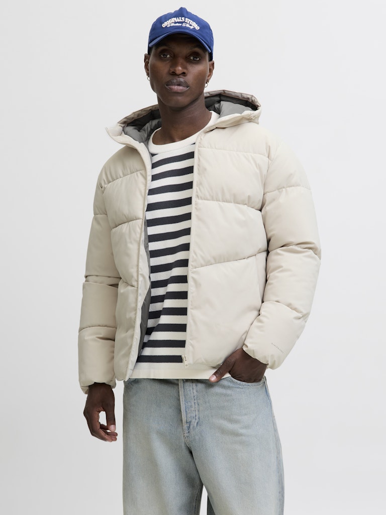 Person wearing a beige puffer jacket, striped shirt, and blue cap on a white background