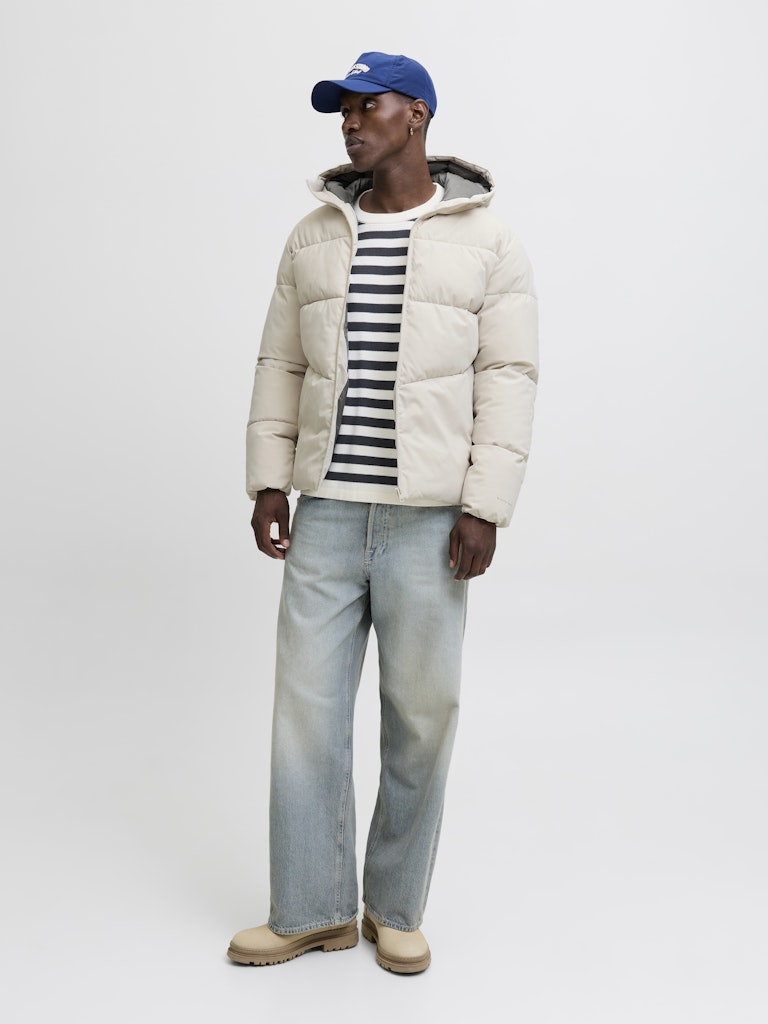 Person wearing a beige puffer jacket, striped shirt, and blue jeans on a white background