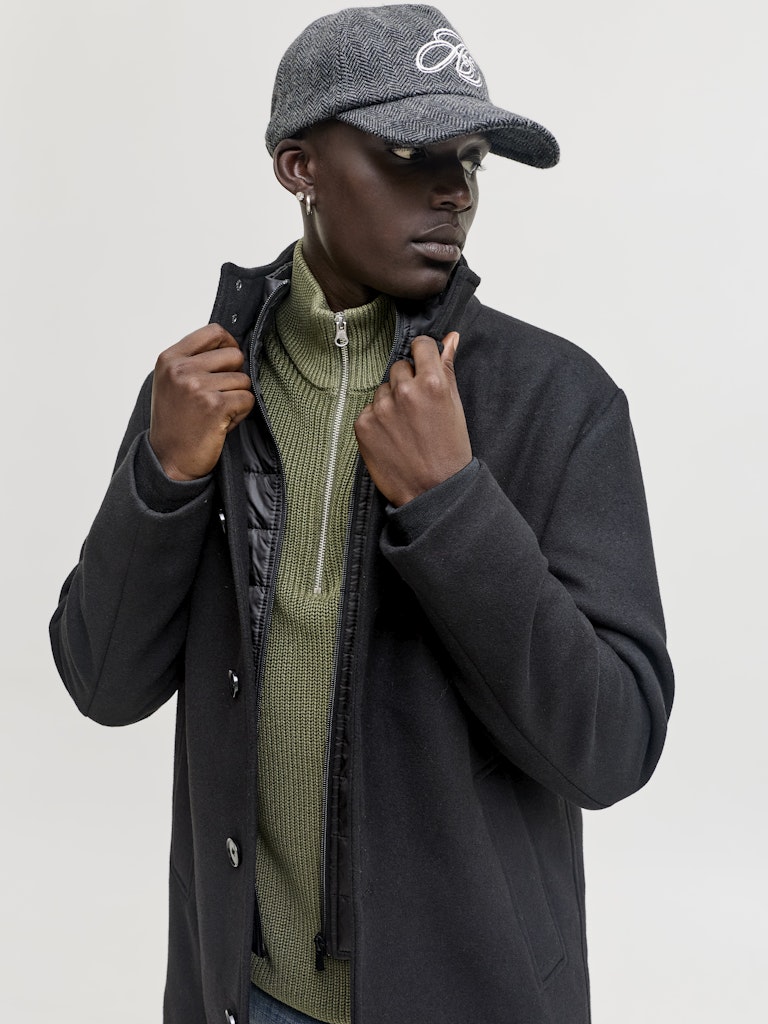 Man wearing a dark coat, green sweater, and gray cap on a light gray background