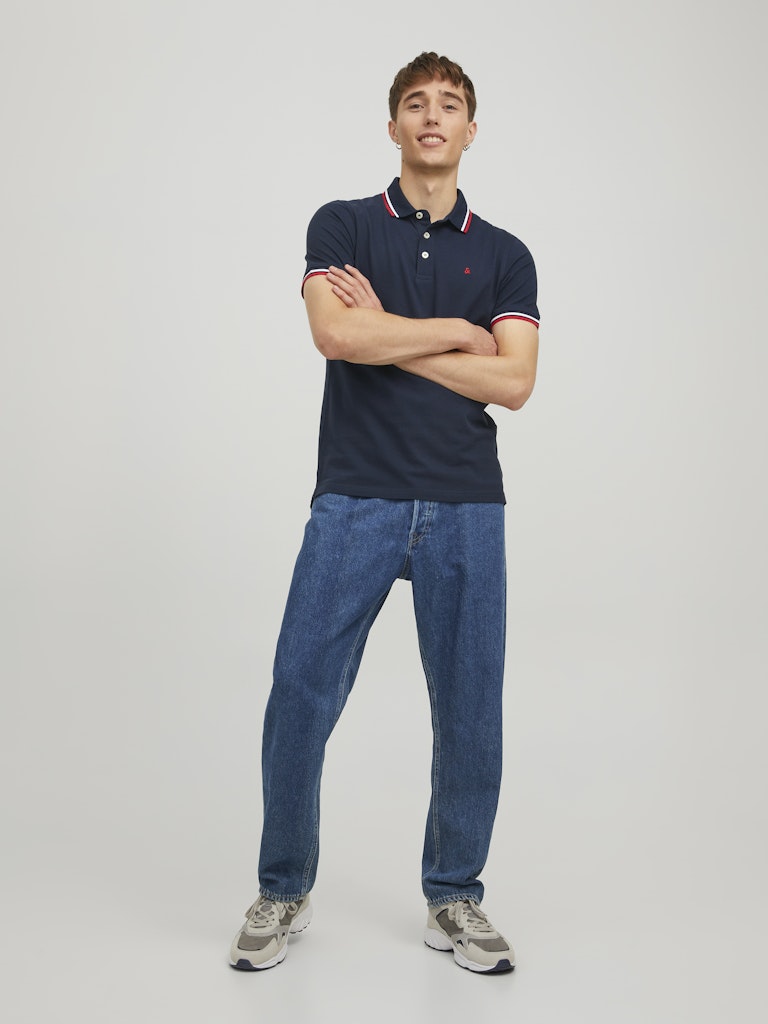 Man wearing a navy polo shirt and blue jeans standing against a plain background