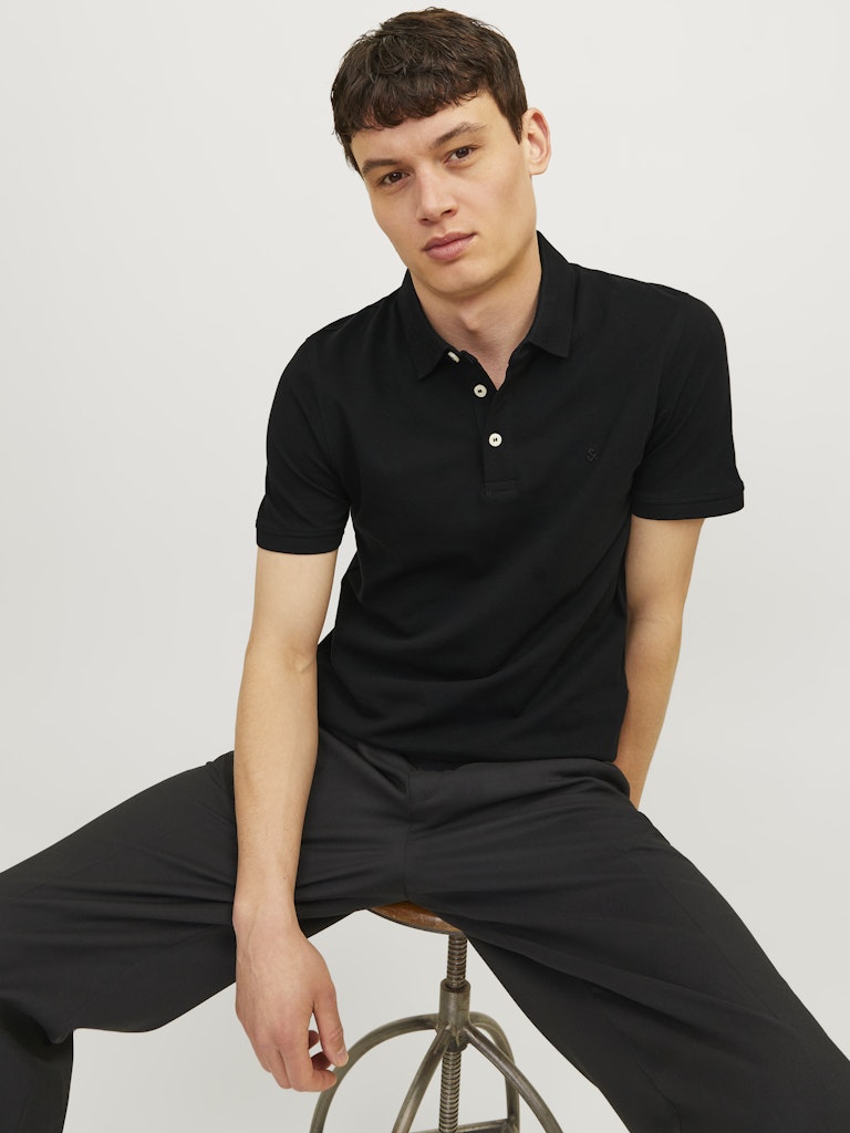 Man wearing a black polo shirt and pants sitting on a stool against a plain background