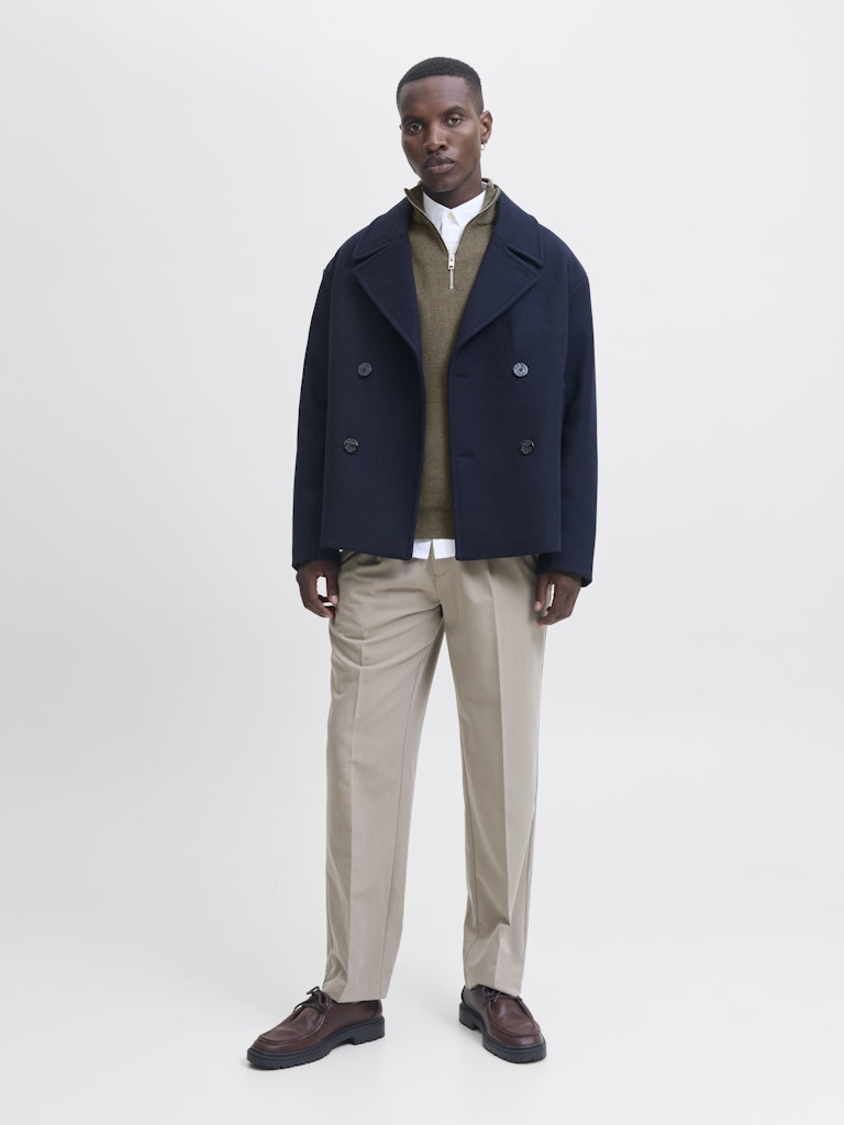 Man wearing a navy peacoat, brown sweater, beige pants, and brown shoes on a white background