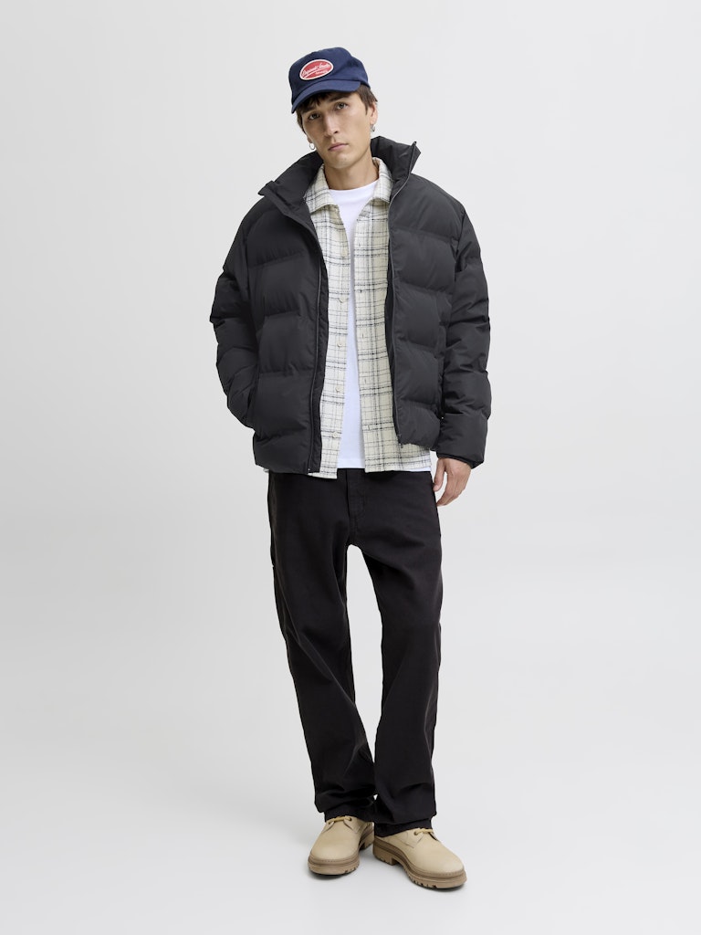 Person wearing a black puffer jacket, plaid shirt, black pants, and beige shoes on a white background