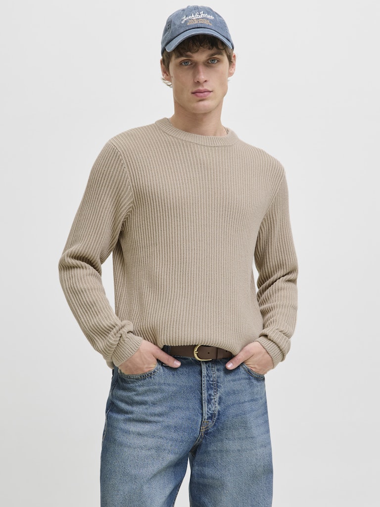 Man wearing a beige sweater, blue jeans, and a blue cap on a light gray background