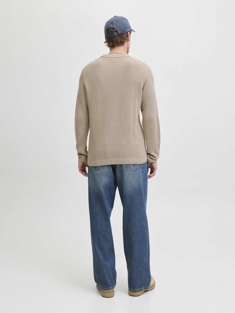 Person wearing a beige ribbed sweater and blue jeans standing against a white background