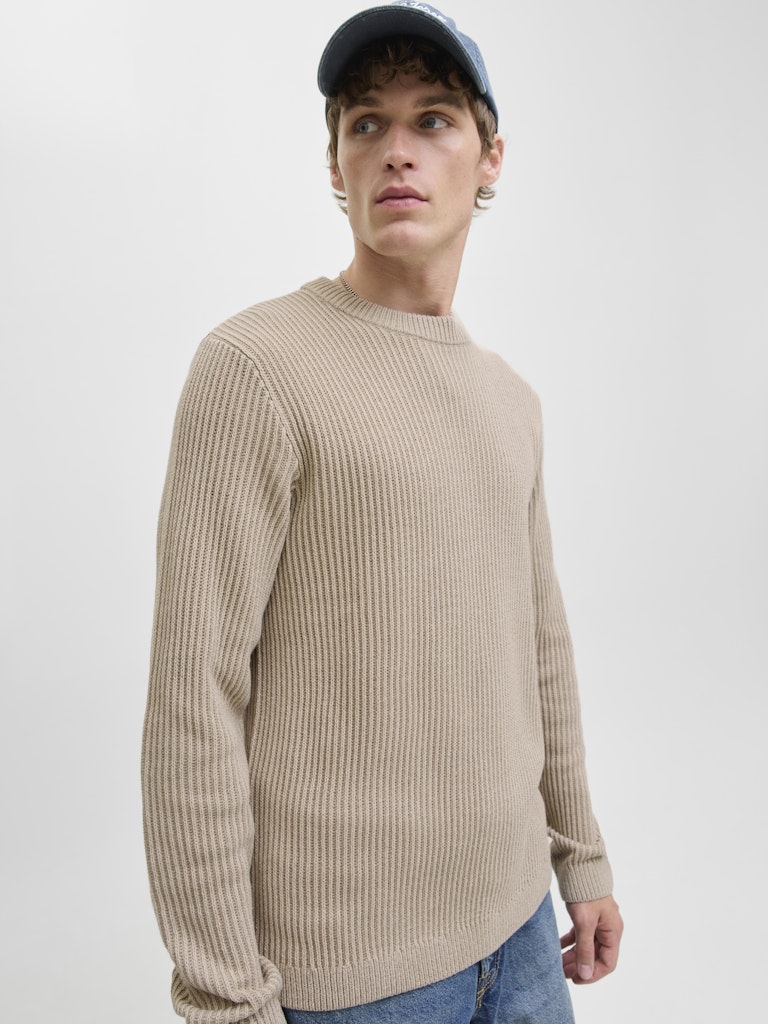 Man wearing a beige ribbed sweater and blue jeans against a plain background