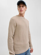 Man wearing a beige ribbed sweater and blue jeans against a plain background