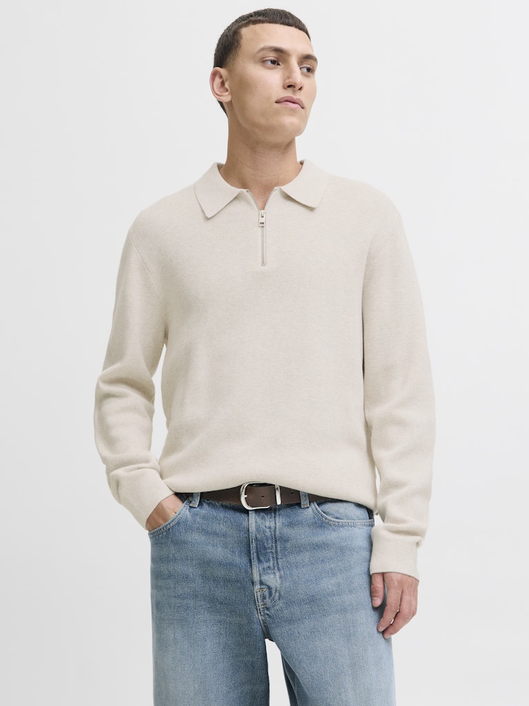 Man wearing a beige sweater and light blue jeans on a white background