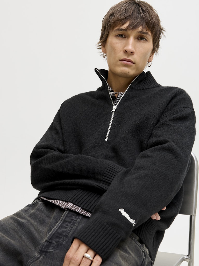 Person wearing a black half-zip sweater with a white brand logo on a light gray background