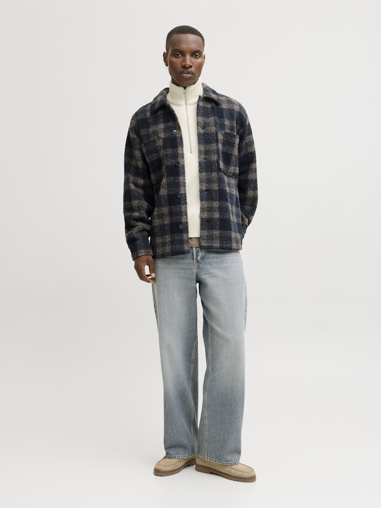 Man wearing a plaid shirt, white sweater, and jeans on a plain background